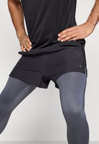 Juodos atletinės trumpos kelnės dėvimos ant pilkų tamprių, pagamintos iš lengvos medžiagos. Trumpikės turi šoninę skylę ir mažą logotipą apačioje.