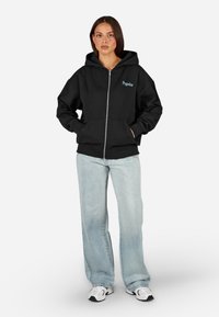 Čierna mikina na zips s predným vreckom a rebrovanými manžetami, so tyrkysovým logom. Skombinované so svetlomodrými širokými džínsami a teniskami.