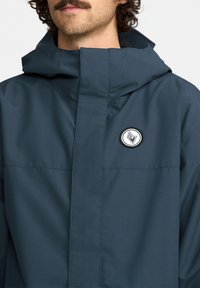 Ciemnoniebieska wodoodporna kurtka z wysokim kołnierzem, zamek błyskawiczny i naszywka z logo. Gładka tekstura i minimalistyczny design. Posiada kaptur.