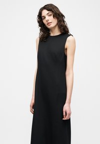 Γυναίκα με σγουρά μαλλιά, που φοράει μαύρο αμάνικο φόρεμα, στέκεται μπροστά σε απλό λευκό φόντο, κοιτάζοντας μπροστά με χαλαρή έκφραση.