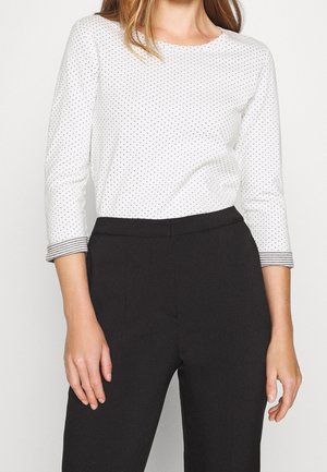 Kobieta w białej koszuli z długim rękawem z małymi czarnymi kropkami i paskowanymi mankietami, w połączeniu z wysokim stanem czarnych spodni.