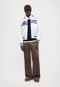Man in een bruine pet, lichtblauwe bomberjack met paarse graphic, witte shirt, marineblauwe sweater, bruine wijde broek en bruine met patroon schoenen.