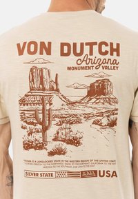 Camiseta beige con un diseño gráfico marrón de Monument Valley, cactus y el texto "VON DUTCH Arizona", con detalles paisajísticos notables.