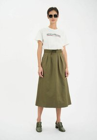 T-shirt blanc avec un texte rouge et bleu, jupe midi plissée olive, et chaussures à talons blocs vertes. Modèle portant des lunettes de soleil noires.