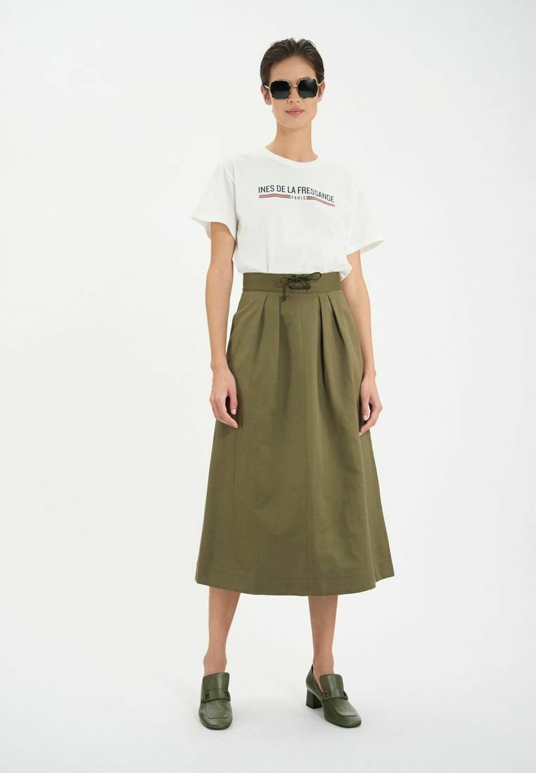 T-shirt blanc avec un texte rouge et bleu, jupe midi plissée olive, et chaussures à talons blocs vertes. Modèle portant des lunettes de soleil noires.