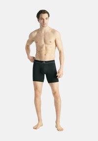 Jeune homme debout pieds nus, portant un boxer noir mi-cuisse avec un logo sur la ceinture, sur un fond clair uni.