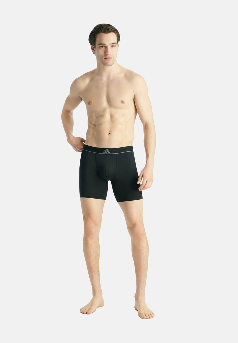 Jeune homme debout pieds nus, portant un boxer noir mi-cuisse avec un logo sur la ceinture, sur un fond clair uni.