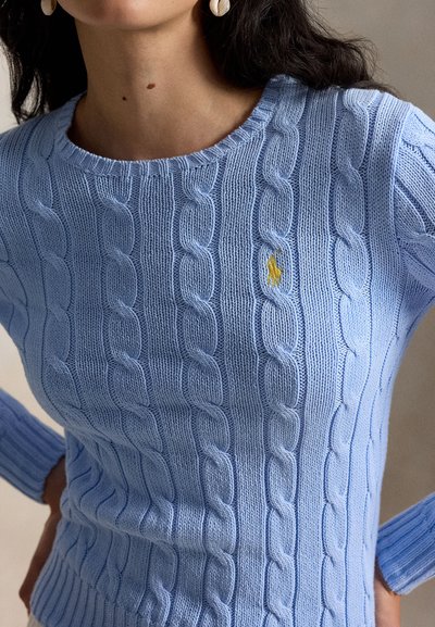 Vrouw draagt een lichtblauwe kabelgebreide trui met een klein geel geborduurd logo op de borst en oorbellen in de vorm van een schelp.