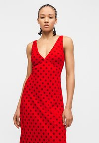 Γυναίκα με πλεξούδες που φοράει ένα κόκκινο φόρεμα χωρίς μανίκια με σκούρες πουά, στέκεται μπροστά σε απλό ανοιχτό φόντο.