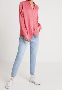 Chemise en lin rose à boutons avec col, poche avant et fentes sur les côtés, portée avec un jean skinny bleu clair et des baskets montantes blanches.