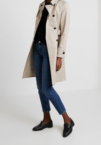 Beige trenchcoat med dubbelknapppning, svarta knappar och bälte i midjan, tillsammans med mörkblå jeans och svarta loafers.