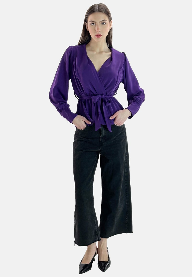 Blouse violette à manches longues et taille nouée, associée à un jean noir à jambe large et des escarpins noirs à bout pointu. Texture du tissu lisse.
