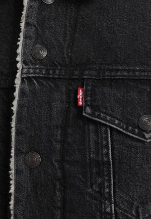 Sort denimjakke med sherpa-foring, metalknapper og et rødt Levi's-brandmærke på lommeflappen.
