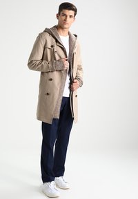Gabardina beige con capucha de punto, botones de doble botonadura, interior texturizado, llevada sobre una camisa blanca y pantalones azul marino, combinada con zapatillas blancas.