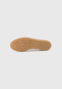 Semelle en caoutchouc de couleur beige avec un motif de grip texturé. Présente des accents circulaires et hexagonaux ; marquage en relief sur le côté.