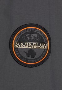 Kuvioitu pyöreä merkki tummanharmaassa kankaassa, jossa on musta ja oranssi teksti, maapallon kuvio ja Antarktiksen ääriviivat keskellä.