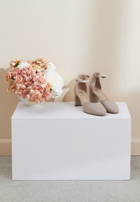 Beżowe szpilki na kostkę umieszczone na białym postumencie, z kwadratowym noskiem i blokowym obcasem. Obok znajduje się wazon z różowymi i białymi kwiatami.