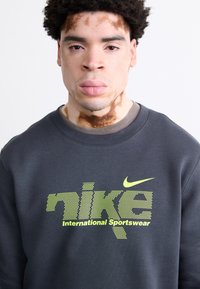 Σκούρο γκρι φούτερ από βαμβακερό μείγμα, με ένα λογότυπο και κείμενο "nike International Sportswear" σε κίτρινο-πράσινο χρώμα μπροστά.