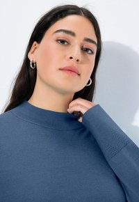 Modrý svetr s vysokým límcem a texturovanými detaily na rukávech, vyrobený ze měkkého pleteného materiálu. Modelka se dotýká výstřihu, na bílém pozadí.