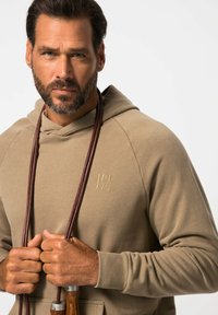 Felpa beige con cappuccio, con una finitura testurizzata e un logo in rilievo. Presenta un cordino e una cintura in pelle marrone tenuta tra le mani.