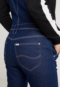 Gros plan d'une personne portant une salopette en denim bleu foncé avec une poche arrière visible et des coutures blanches, main reposant dans la poche latérale.