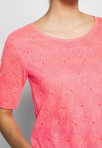 Femme portant un haut corail à manches courtes avec un motif floral géométrique texturé et un col rond.