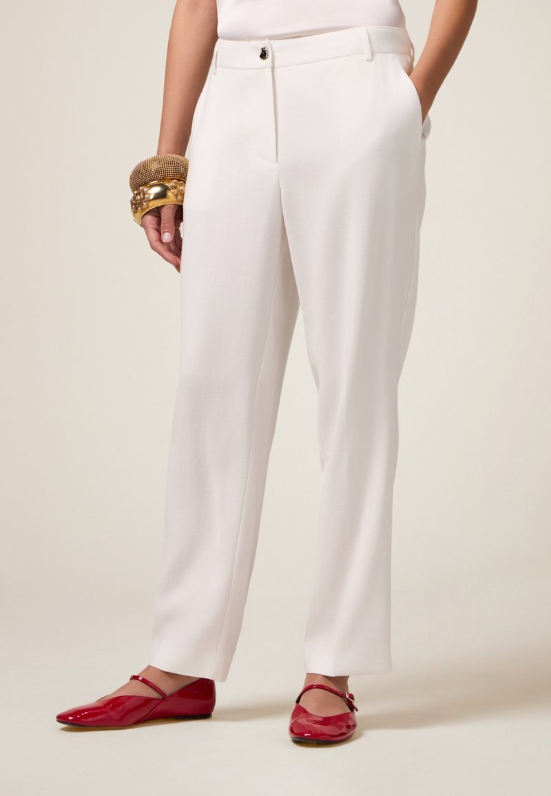 Pantalon blanc ajusté avec une coupe droite, présentant des poches latérales et une fermeture à bouton. Associé à des ballerines rouges et des bracelets en or.
