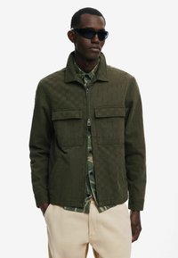 Olivengrøn jakke med tekstureret mønster, lynlås lukning og to forlommer, lagd فوق en camouflage skjorte, parret med beige bukser.