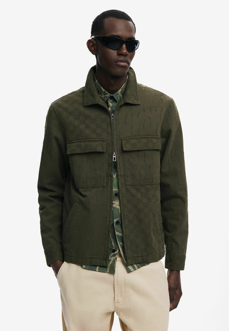 Olivengrøn jakke med tekstureret mønster, lynlås lukning og to forlommer, lagd فوق en camouflage skjorte, parret med beige bukser.
