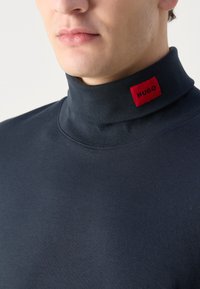 Pullover a collo alto di un blu navy scuro con una consistenza liscia e un prominente etichetta rossa "HUGO" cucita al colletto. Visione ravvicinata della parte superiore del petto.