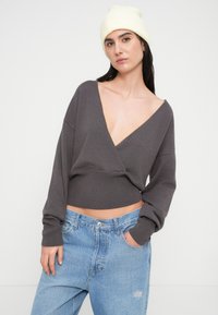 Γκρι κοντό πουλόβερ με ντεκολτέ σε σχήμα V και φαρδιά μανίκια, συνδυασμένο με ανοιχτό μπλε τζιν. Το μοντέλο φοράει λευκό πλεκτό σκουφάκι.