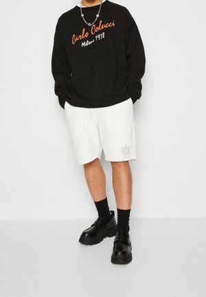 Svart sweatshirt med orange och vit text; vita shorts med två fickor och broderad logotyp; svarta strumpor och kraftiga svarta skor.