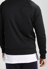 Černá mikina s přiléhavým střihem, dlouhými rukávy, žebrovanými manžetami a lemem a hladkým, mírně texturovaným materiálem. Obsahuje kulatý výstřih.