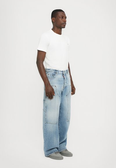 Vitt kortärmat t-shirt som matchas med oversized ljusblå denimjeans med fickor och blekning. Modellen har på sig grå skor.