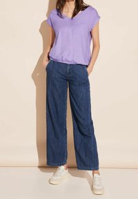 Haut violet à manches courtes avec un ourlet froncé, associé à un jean en denim bleu à jambes larges et des baskets blanches avec des accents beiges.