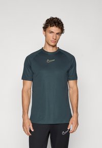 Tumši teāla Nike sporta krekls ar īsām piedurknēm, apaļu dekoltē un nelielu dzeltenu logotipu uz krūtīm. Izgatavots no gludas, vieglas auduma.