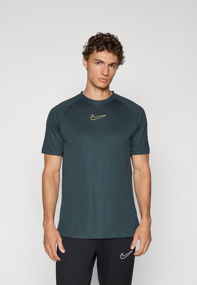 Tumši teāla Nike sporta krekls ar īsām piedurknēm, apaļu dekoltē un nelielu dzeltenu logotipu uz krūtīm. Izgatavots no gludas, vieglas auduma.