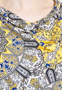 Tissu à motifs présentant des designs complexes en noir, jaune et bleu. Texture douce avec un col drapé. Motifs floraux et géométriques notables.
