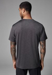 Camiseta deportiva de manga corta en gris oscuro con una tela texturizada y un cuello redondo, que presenta un corte relajado y tatuajes visibles en el brazo.