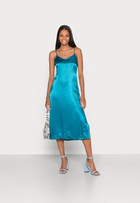 Vestido de cetim verde-água com alças finas e comprimento midi. Combinado com sandálias de salto bloco azuis e uma mala com estampado de zebra preto e branco.