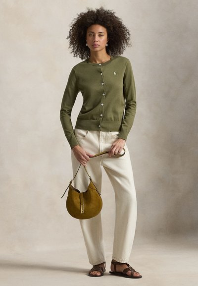 Πράσινη ζακέτα σε χρώμα ελιάς με κουμπιά, κρεμ παντελόνι και καφέ δερμάτινα σανδάλια. Αξεσουάρ περιλαμβάνουν μία στρογγυλή τσάντα σουέντ σε χρώμα μουσταρδί.