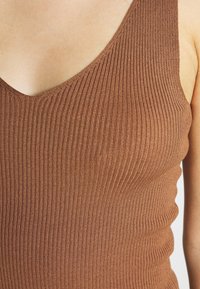 Gros plan sur une femme portant un haut marron sans manches à côtes avec un décolleté en V, montrant son épaule et le haut de sa poitrine.