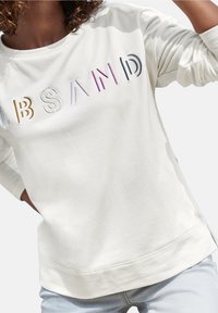 Sudadera crema con texto bordado en relieve y colorido que dice "ABSAND" en la parte frontal. Fabricada en un material suave con un corte relajado y puños acanalados.