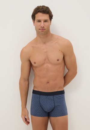 Oberkörperfreier Mann mit kurzen lockigen braunen Haaren, der blaue gemusterte Boxershorts trägt und vor einem schlichten hellen Hintergrund steht.