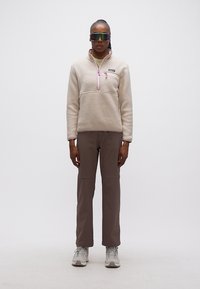 Γυναίκα που στέκεται φορώντας μπεζ φλις μπουφάν, καφέ παντελόνι, ανοιχτό γκρι παπούτσια πεζοπορίας και μεγάλα αντανακλαστικά γυαλιά ηλίου μπροστά σε απλό φόντο.