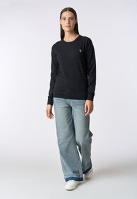 Maglione nero a maniche lunghe in materiale morbido con scollo rotondo e un piccolo logo sul petto, abbinato a jeans larghi azzurri.