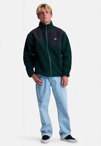 Zielona kurtka z polaru z czarnymi panelami, wysokim kołnierzem, przednim zamkiem błyskawicznym, z logo na piersi. Nosi się ją z jasnoniebieskimi dżinsami i czarnymi butami.