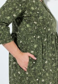 Abito floreale verde con maniche a tre quarti, caratterizzato da una vita arricciata, tasche laterali e un motivo in tessuto testurizzato.