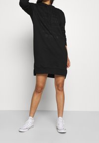 Osoba v černých dlouhých šatech s kapucí a tištěným textem, stojící na jednoduchém pozadí, obuta v bílých vysokých teniskách.