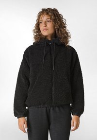 Giacca in pile nera con cappuccio, dotata di chiusura a zip, polsini elasticizzati e superficie texture. Indossata con pantaloni grigio scuro.